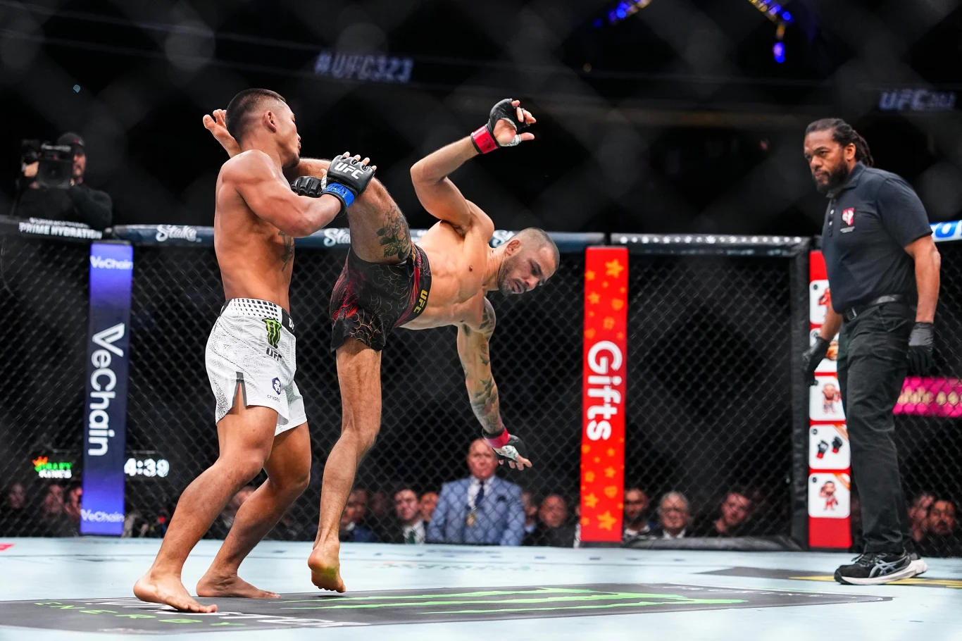 Dwóch zawodników MMA walczy w oktagonie, jeden z nich wykonuje efektowny kopnięcie obrotowe w powietrzu, sędzia obserwuje pojedynek z bliska, a w tle widoczna jest publiczność i reklamy.