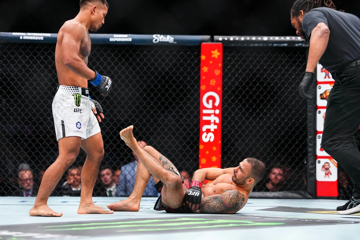 Dwóch zawodników walczy w klatce UFC, jeden z nich leży na ziemi, podczas gdy drugi stoi obok niego. W tle widoczny jest sędzia, fragmenty reklamy oraz publiczność.