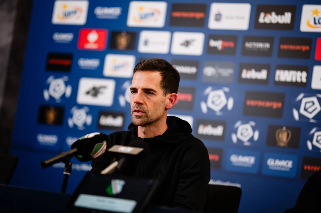 Inaki Astiz Mężczyzna w czarnej bluzie siedzący przy stole konferencyjnym z mikrofonami, w tle ścianka z logotypami sponsorów i organizatorów wydarzenia sportowego.