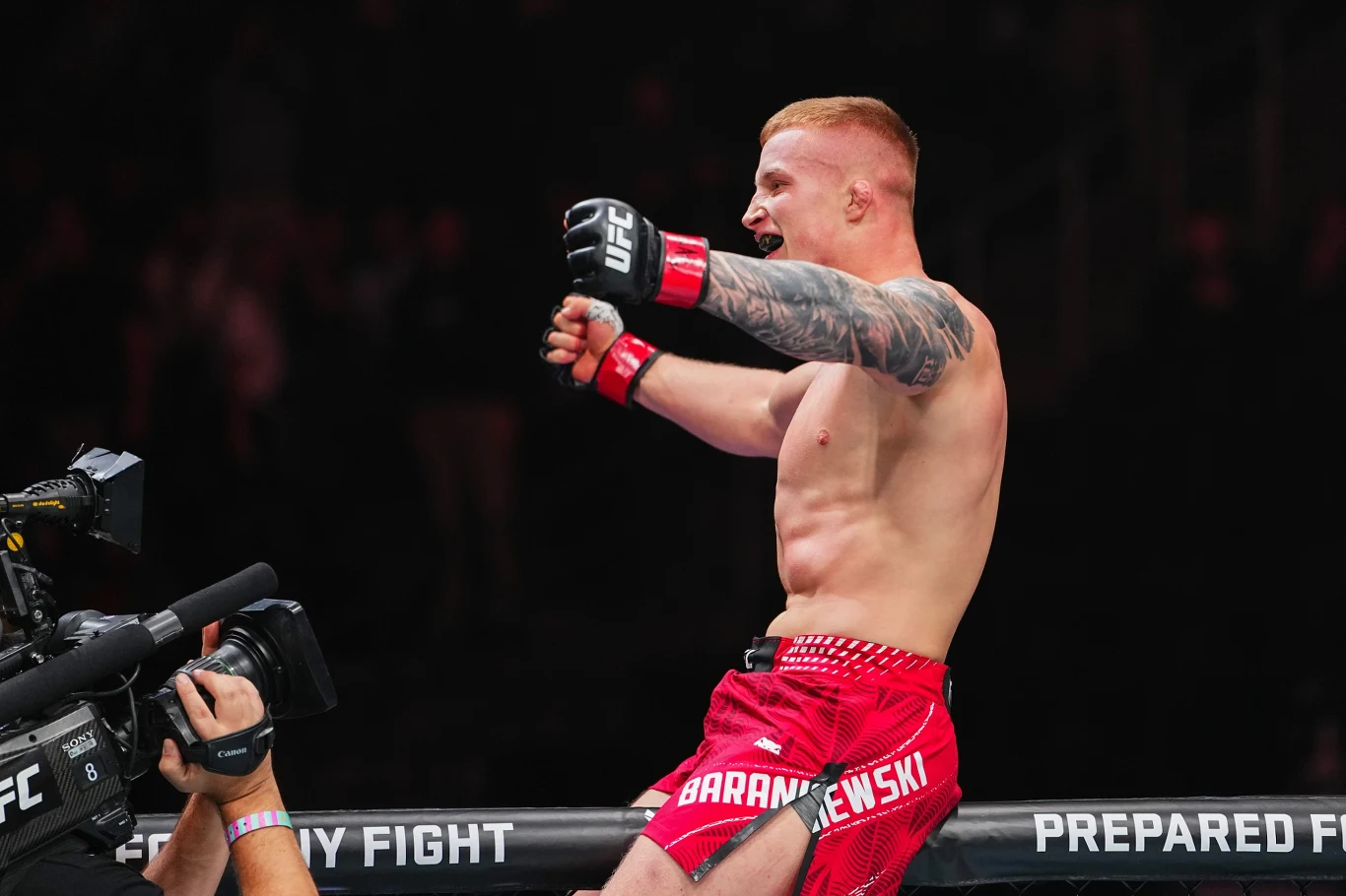 Iwo Baraniewski Zawodnik MMA stojący w oktagonie wyraża radość po zwycięstwie, z rękami uniesionymi w geście triumfu. Mężczyzna ubrany w czerwone spodenki z widocznym tatuażem na ramieniu, w tle widać kamerzystę filmującego wydarzenie.