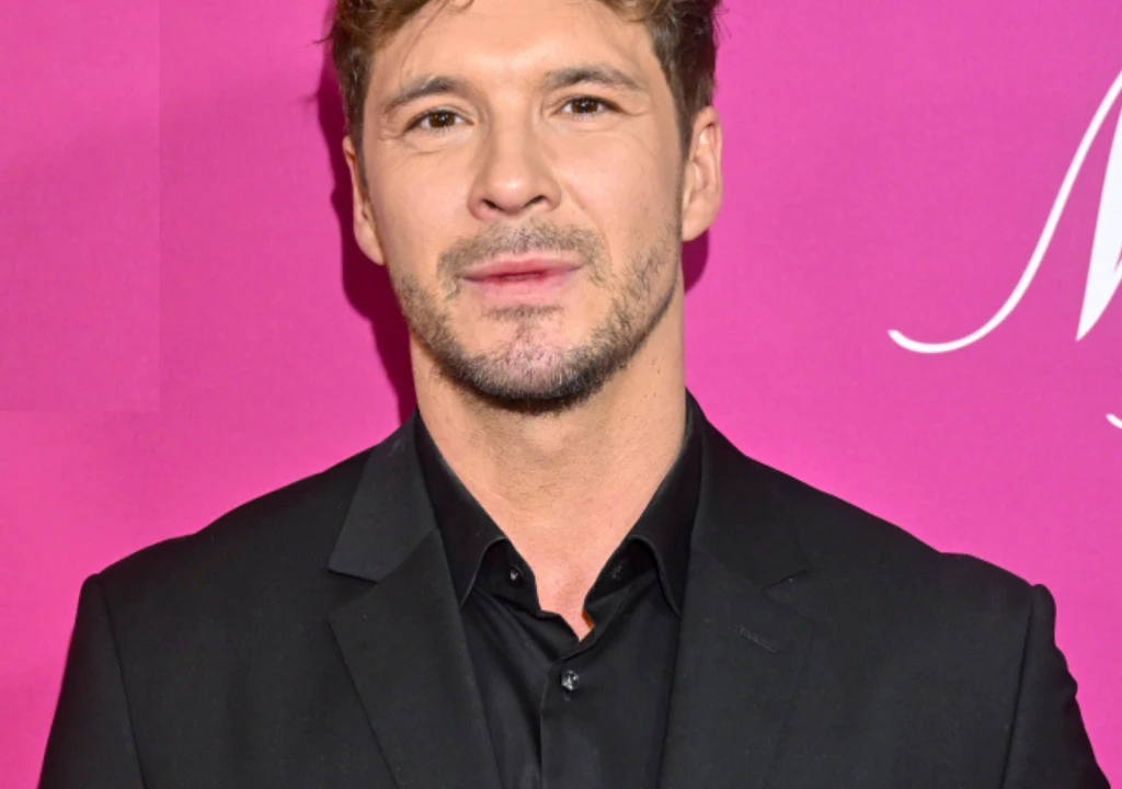 Mikołaj Roznerski Mężczyzna w eleganckim, czarnym garniturze na tle jasnoróżowej, jednolitej ścianki, włosy ułożone w luźną fryzurę, lekki zarost, wyraz twarzy neutralny.