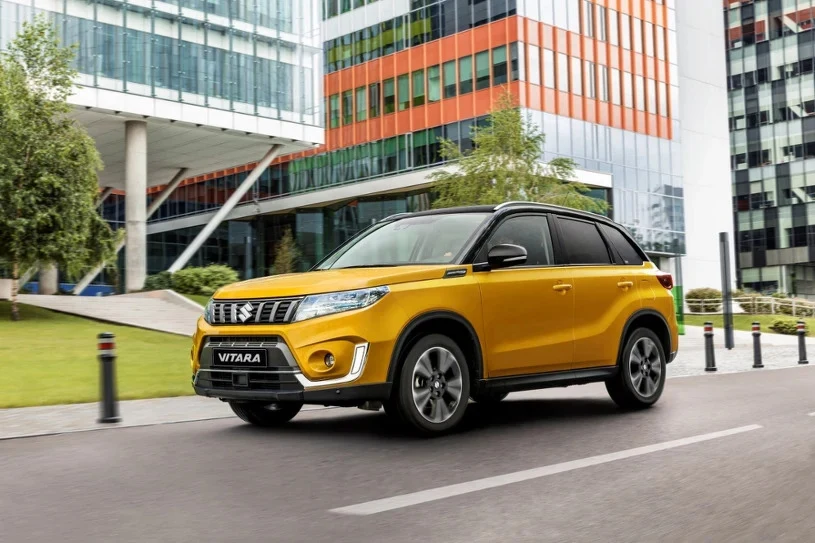 Suzuki Vitara co prawda jest prosta konstrukcyjnie, ale dzięki temu rzadko się psuje Żółto-czarny samochód SUV Suzuki Vitara jadący ulicą przy nowoczesnych szklanych budynkach biurowych, w tle zieleń oraz elementy miejskiej infrastruktury.