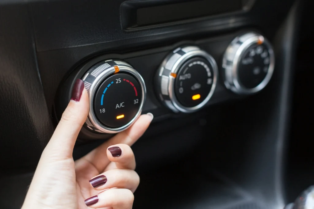 Kobieta z lakierowanymi paznokciami ustawia pokrętło klimatyzacji w samochodzie, widoczne są także dwa inne pokrętła służące do regulacji nawiewu i temperatury, panele mają podświetlone wskaźniki.