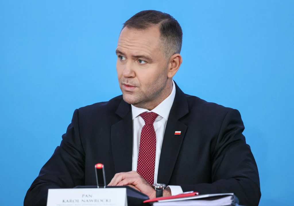 Mężczyzna w garniturze i czerwonym krawacie siedzi przy stole konferencyjnym na tle niebieskiej ściany, przed nim znajduje się mikrofon, dokumenty oraz tabliczka z nazwiskiem.