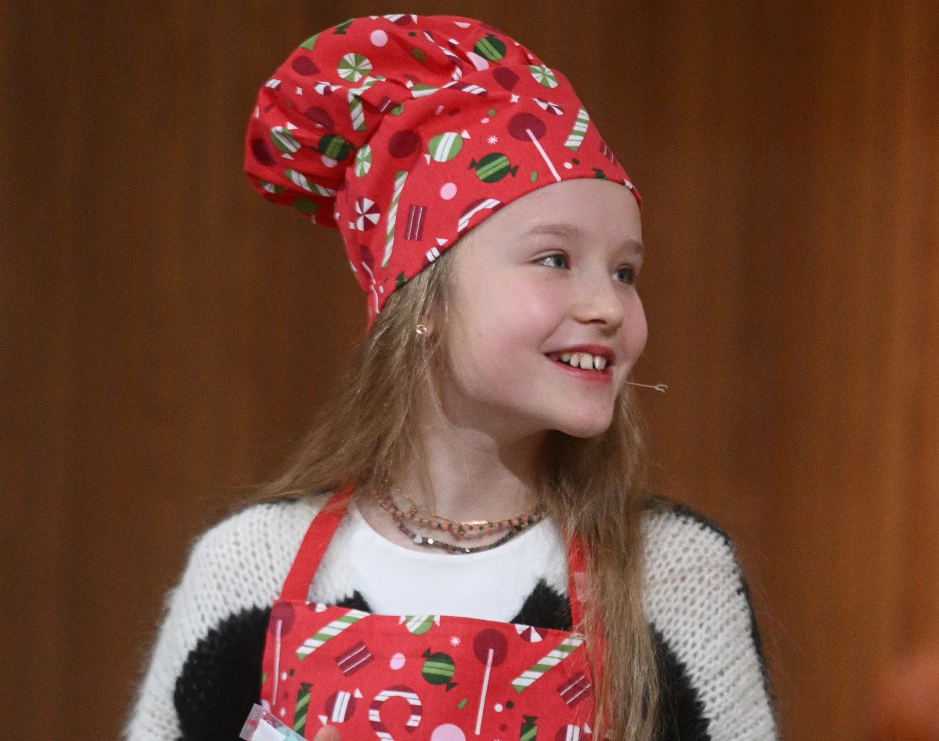 Marianna Kłos reprezentuje Polskę na Eurowizji Junior 2025 Dziewczynka w czerwonej czapce kucharskiej oraz fartuszku w świąteczne wzory, uśmiechnięta, z długimi blond włosami, zwrócona w bok, na tle brązowej ściany.