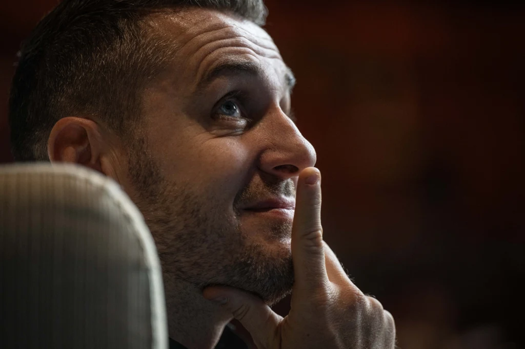 Mark Selby Mężczyzna z krótkim zarostem siedzący na jasnym fotelu, patrzący w górę z uśmiechem i oparciem palca wskazującego o brodę, wyrażający zamyślenie lub zainteresowanie.