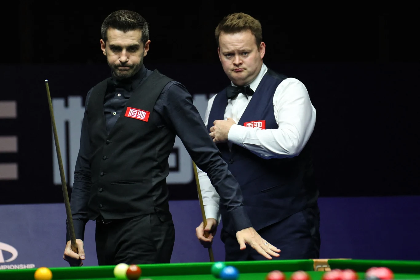 Dwóch mężczyzn w eleganckich kamizelkach i muchach pochylonych nad stołem bilardowym podczas meczu snookera, na pierwszym planie kolorowe bile.