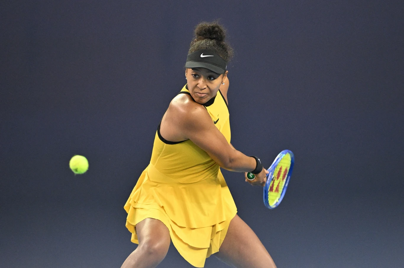 Naomi Osaka Tenisistka w żółtej sukience wykonuje zamach na kort tenisowy, koncentrując się na zbliżającej się piłce, na tle jednolitego, ciemnego tła.