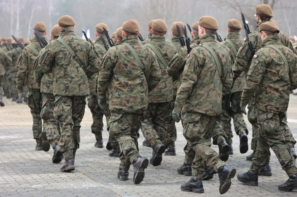 Grupa żołnierzy w mundurach wojskowych maszeruje po placu, wszyscy ubrani są w berety i niosą broń na plecach, widoczny charakterystyczny wzór munduru oraz flagi na ramionach.