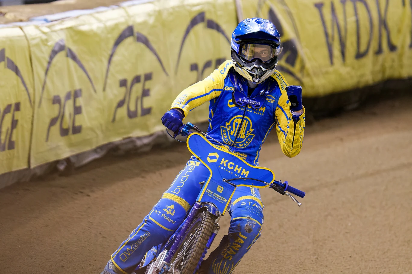 Zawodnik żużlowy w niebiesko-żółtym kombinezonie i kasku wykonuje gest triumfu podczas jazdy na torze, w tle widoczna bariera z reklamami.