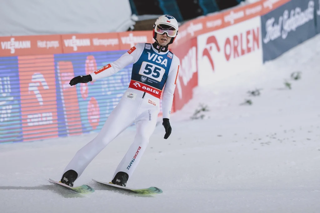 Kamil Stoch Zawodnik w kombinezonie narciarskim ląduje po skoku na nartach podczas zawodów, na tle band z reklamami i śnieżnym podłożem.