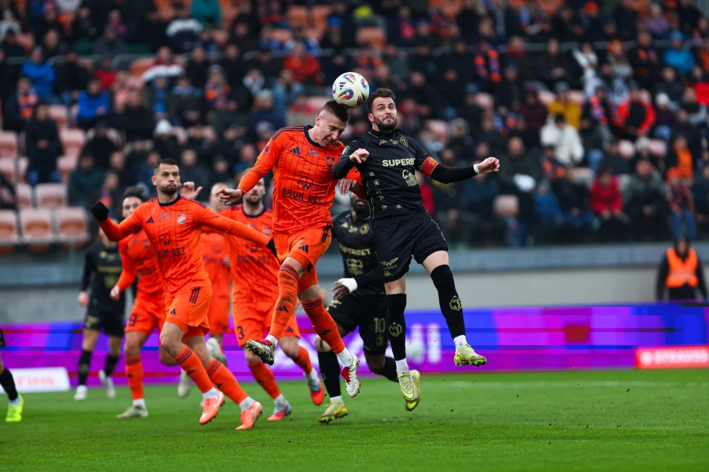 Mecz Bruk-Bet Termalica Nieciecza - Jagiellonia Białystok Zawodnicy dwóch drużyn walczą o piłkę głowami podczas dynamicznej akcji na boisku, w tle widoczna publiczność na trybunach.