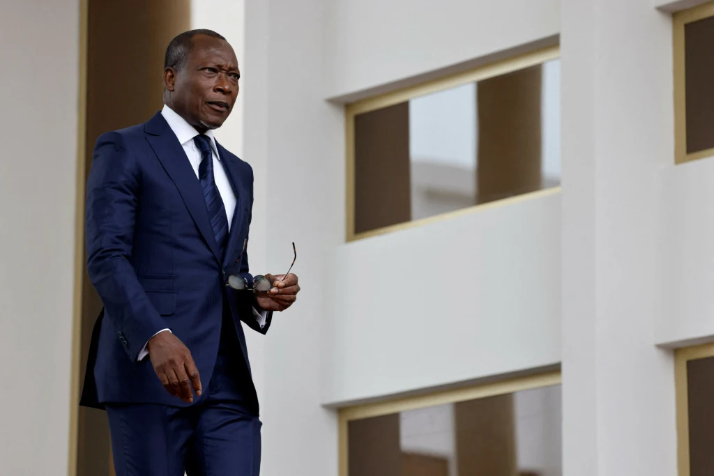 Prezydent Beninu Patrice Talon Mężczyzna w eleganckim, granatowym garniturze stoi na tle jasnych, nowoczesnych budynków, trzymając w jednej ręce okulary.