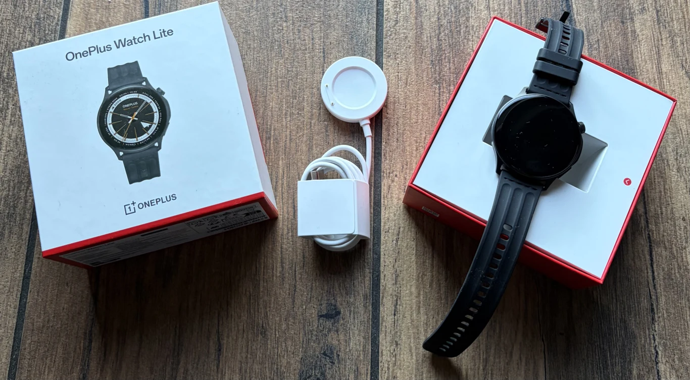 Zawartość opakowania z OnePlus Watch Lite. Zegarek OnePlus Watch Lite z czarnym paskiem ułożony obok swojego oryginalnego opakowania oraz ładowarki na drewnianej powierzchni.