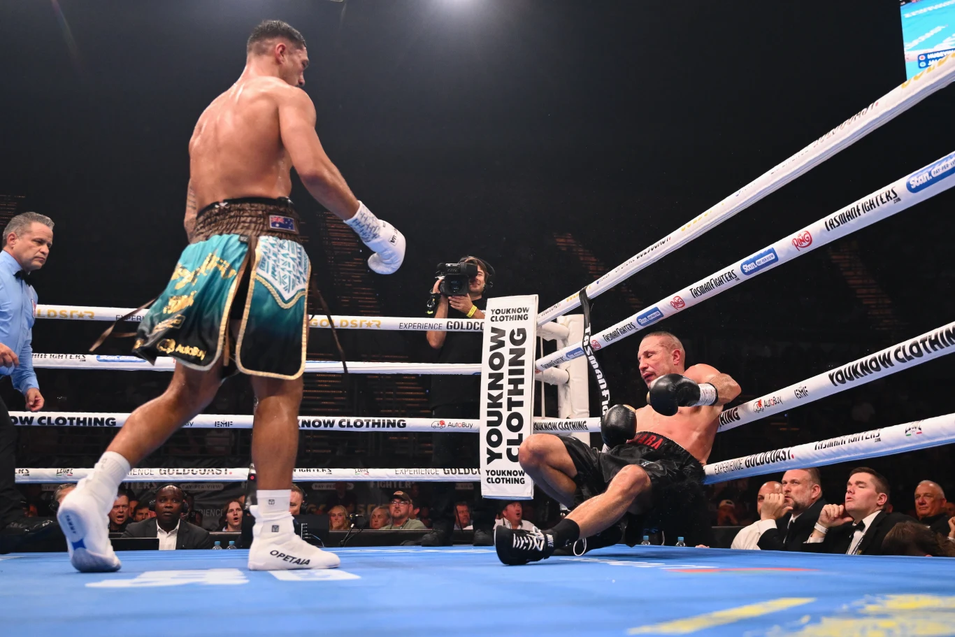 Zawodnik w zielonych spodenkach stoi w ringu, podczas gdy jego przeciwnik w czarnych spodenkach siedzi na podłodze oparty o liny po otrzymanym ciosie, wokół widzowie oraz sędzia.