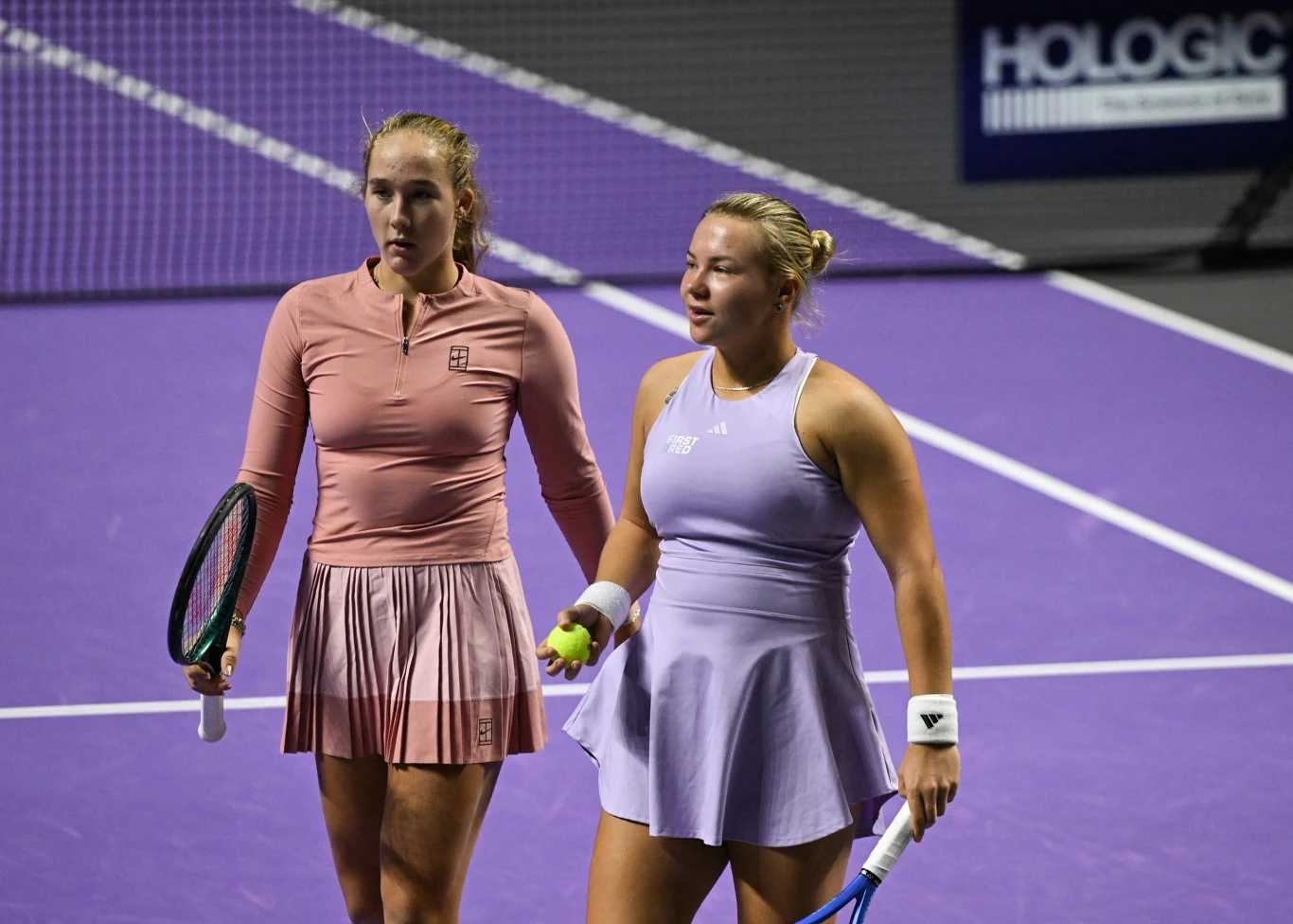 Dwie tenisistki stojące na fioletowym korcie, ubrane w sportowe stroje, trzymające rakiety tenisowe, jedna z piłką w ręce, skupione i gotowe do dalszej gry.