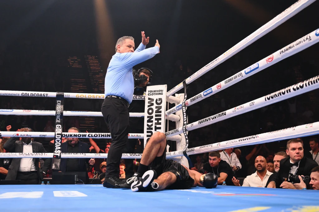 Huseyin Cinkara na deskach Sędzia przerywa walkę bokserską, stojąc nad leżącym na macie zawodnikiem, wokół ringu widoczna jest publiczność oraz ekipa sędziowska i organizacyjna.