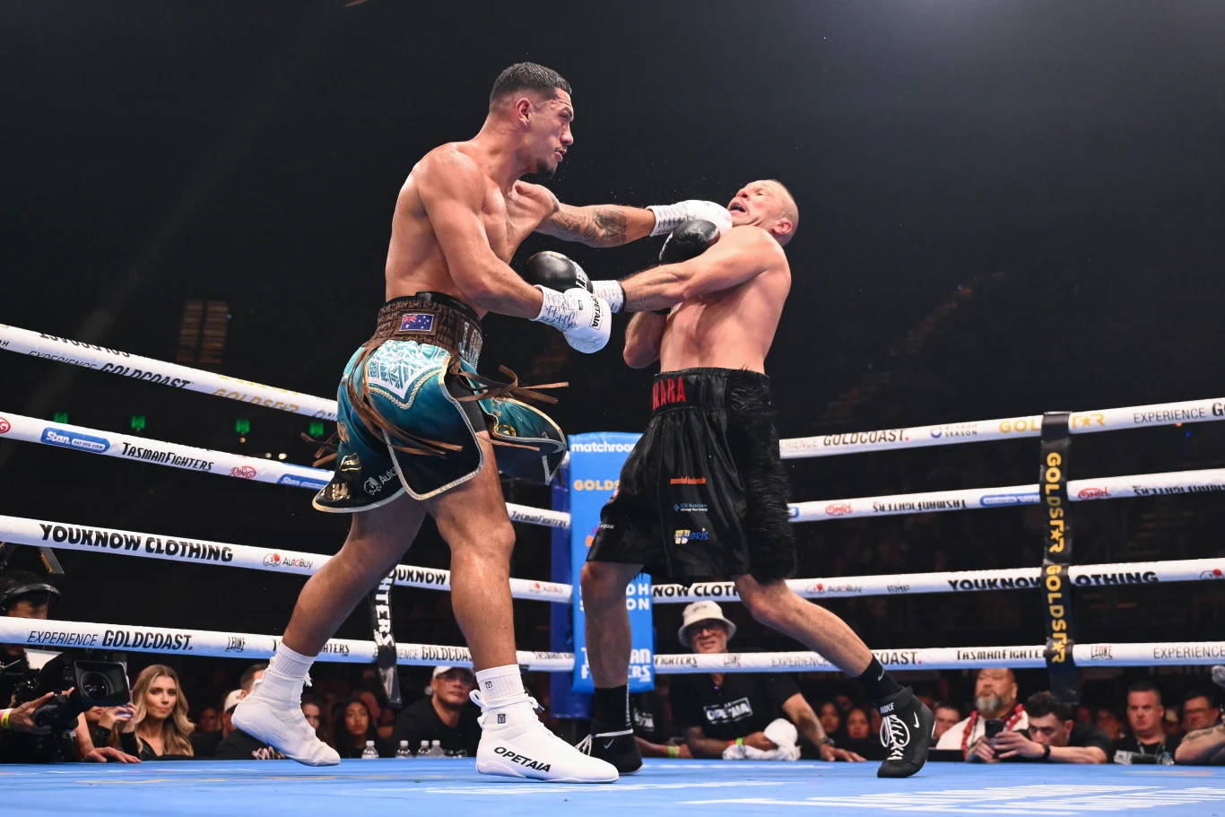 Dwóch bokserów walczy na ringu podczas oficjalnej gali. Zawodnik z lewej zadaje silny cios przeciwnikowi, który cofa się na linach. W tle widać widownię i sędziów obserwujących pojedynek.