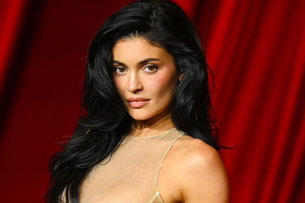 Kylie Jenner Kobieta o długich, czarnych włosach ubrana w przezroczystą bluzkę pozuje na tle czerwonej, marszczonej tkaniny, patrząc prosto w obiektyw aparatu.