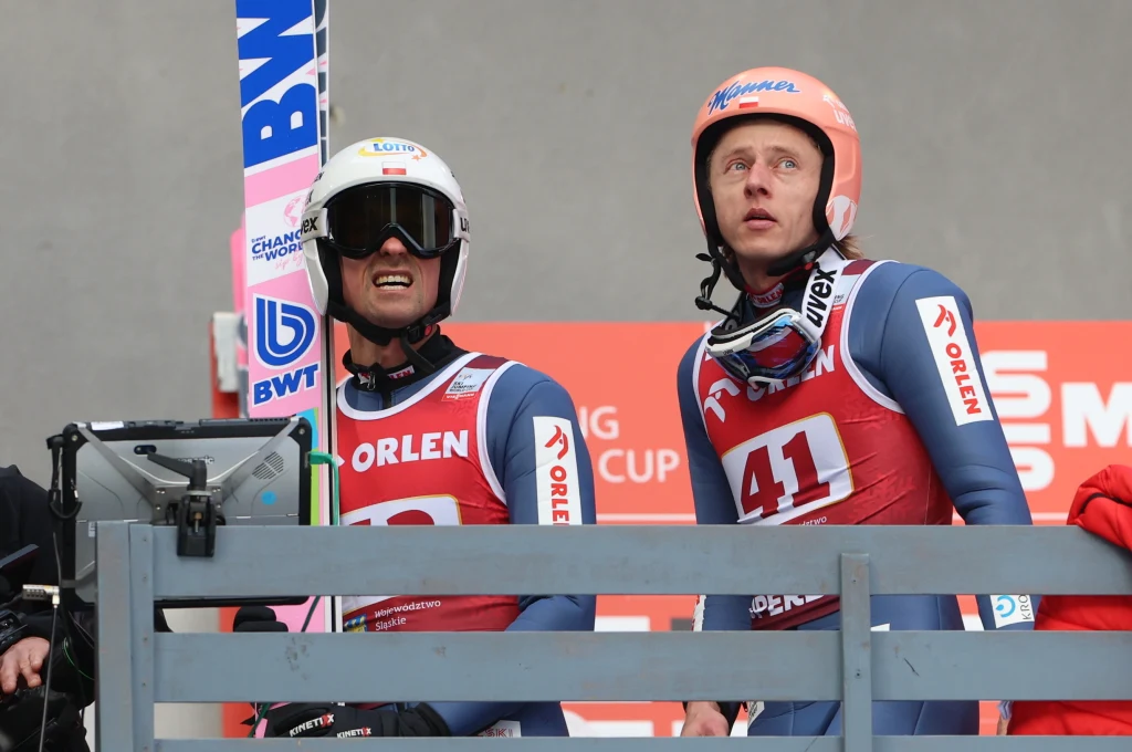 Dawid Kubacki (po prawej) i Piotr Żyła Dwóch skoczków narciarskich w kombinezonach z numerami startowymi i kaskami, stojących przy barierce na tle banerów sponsorskich i ekranu.