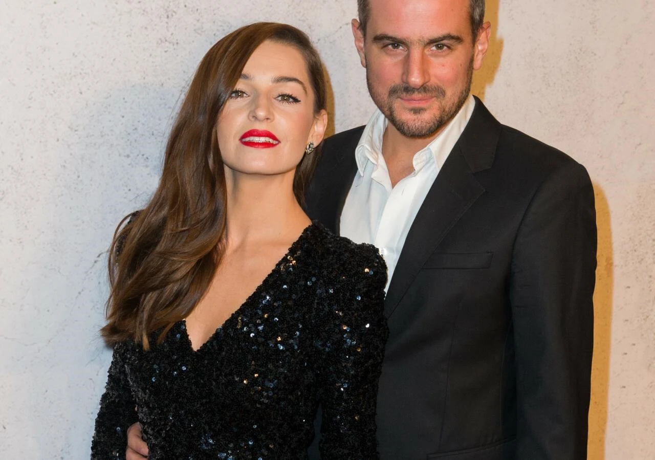 Agnieszka Więdłocha, Antoni Pawlicki Dwie elegancko ubrane osoby stoją razem na tle jasnej ściany, kobieta w czarnej, błyszczącej sukience i mężczyzna w czarnym garniturze z białą koszulą, oboje uśmiechnięci, obejmują się.