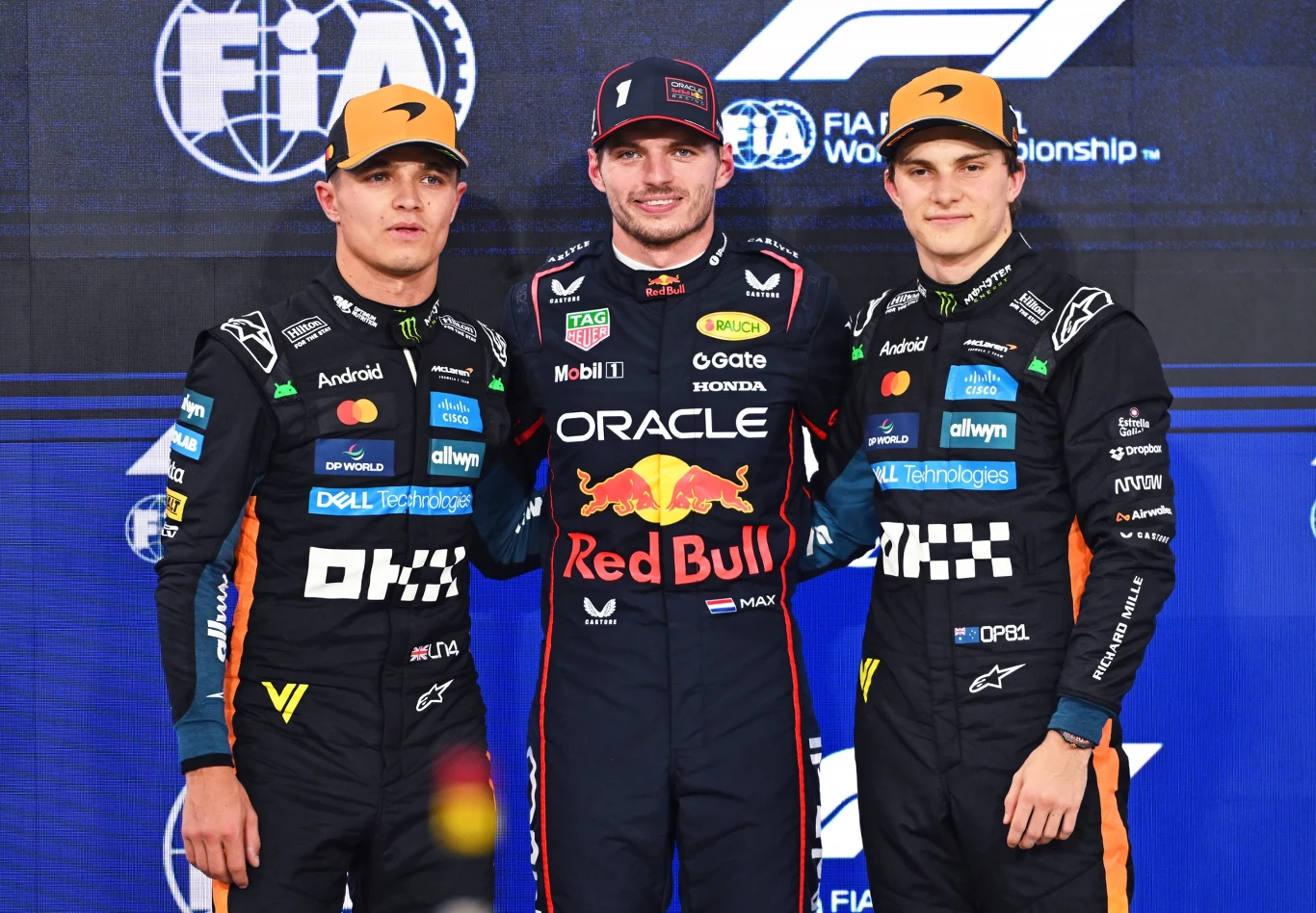 Lando Norris, Max Verstappen, Oscar Piastri Trzech kierowców Formuły 1 pozuje razem na tle ściany z logotypami FIA oraz F1 w swoich oficjalnych kombinezonach zespołowych, dwóch z nich ubranych w pomarańczowe czapki, a centralny zawodnik nosi strój Red Bull Racing.
