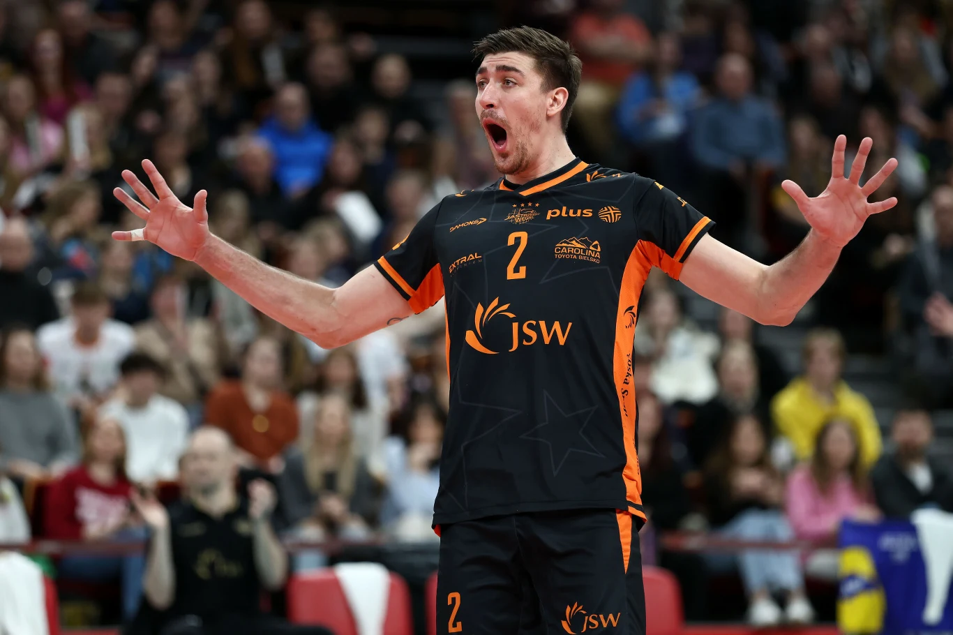 Siatkarz w czarnym stroju z pomarańczowymi wstawkami i numerem 2 gestykuluje szeroko rozchylonymi dłońmi, stoi na tle rozmytej widowni w hali sportowej.