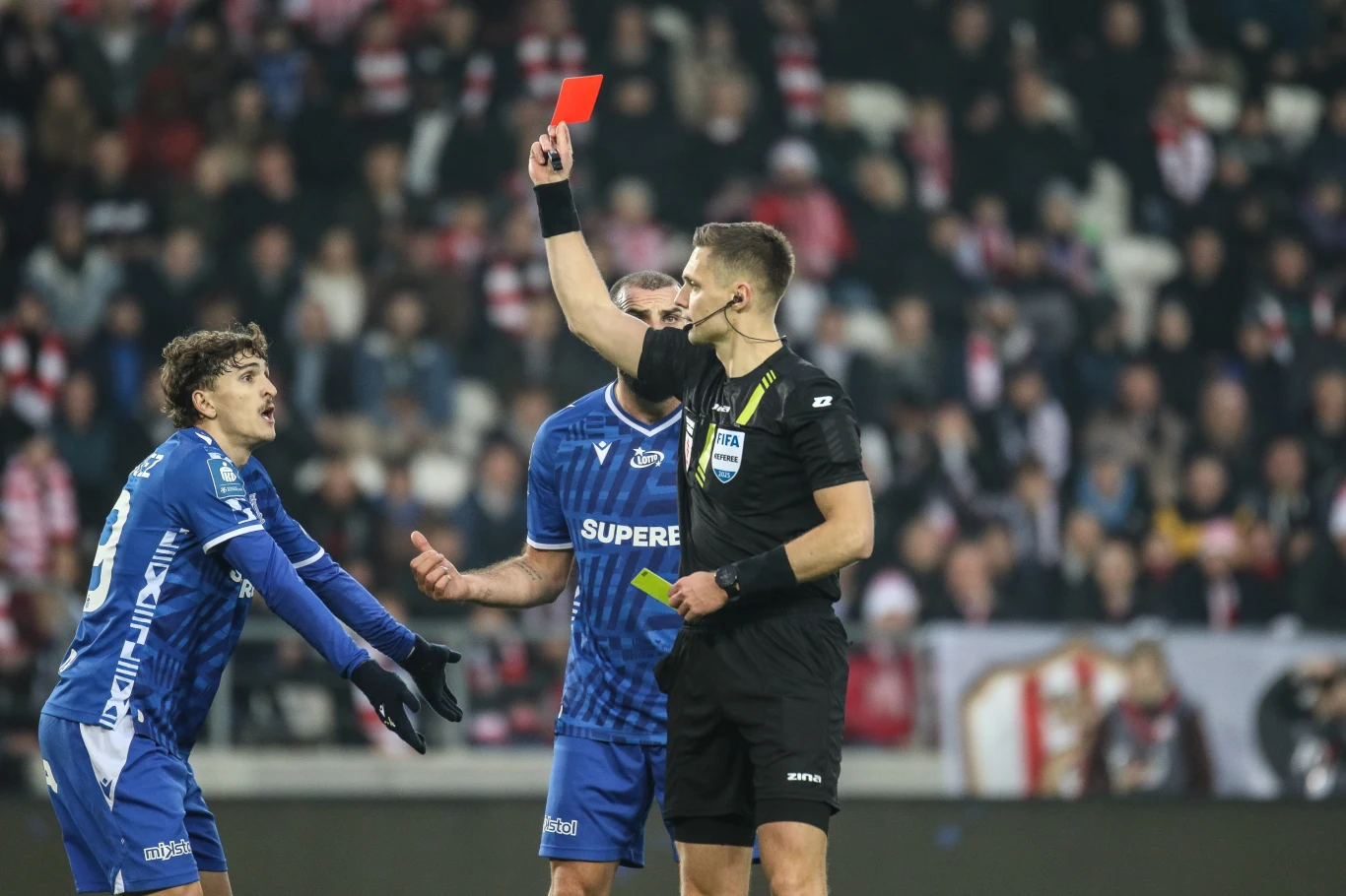 W tej sytuacji sędzia Damian Sylwestrzak nie okazał łaski Sędzia pokazuje czerwoną kartkę piłkarzowi drużyny w niebieskich strojach podczas meczu piłkarskiego, zawodnik gestykuluje zaskoczony, w tle rozmyte postacie kibiców na trybunach.
