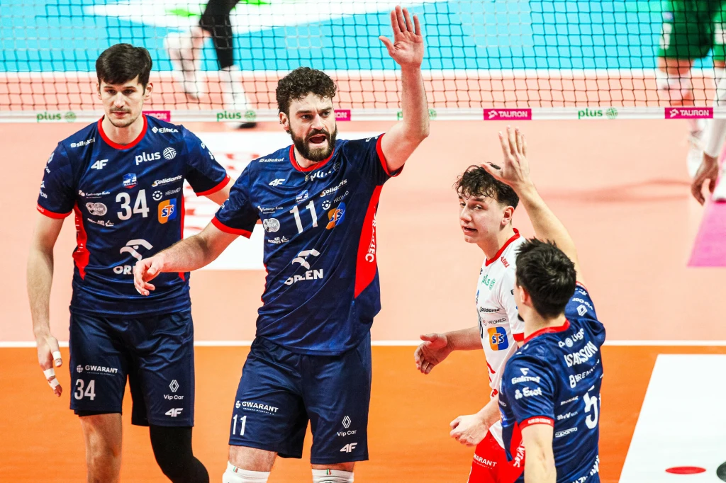 Siatkarze ZAKS-y Kędzierzyn-Koźle. Od lewej Szymon Jakubiszak i Kamil Rychlicki, medaliści ostatnich MŚ Zawodnicy drużyny siatkarskiej w niebieskich i białych strojach świętują zdobycie punktu na boisku, unosząc ręce w geście radości. W tle widoczna siatka oraz fragment publiczności.