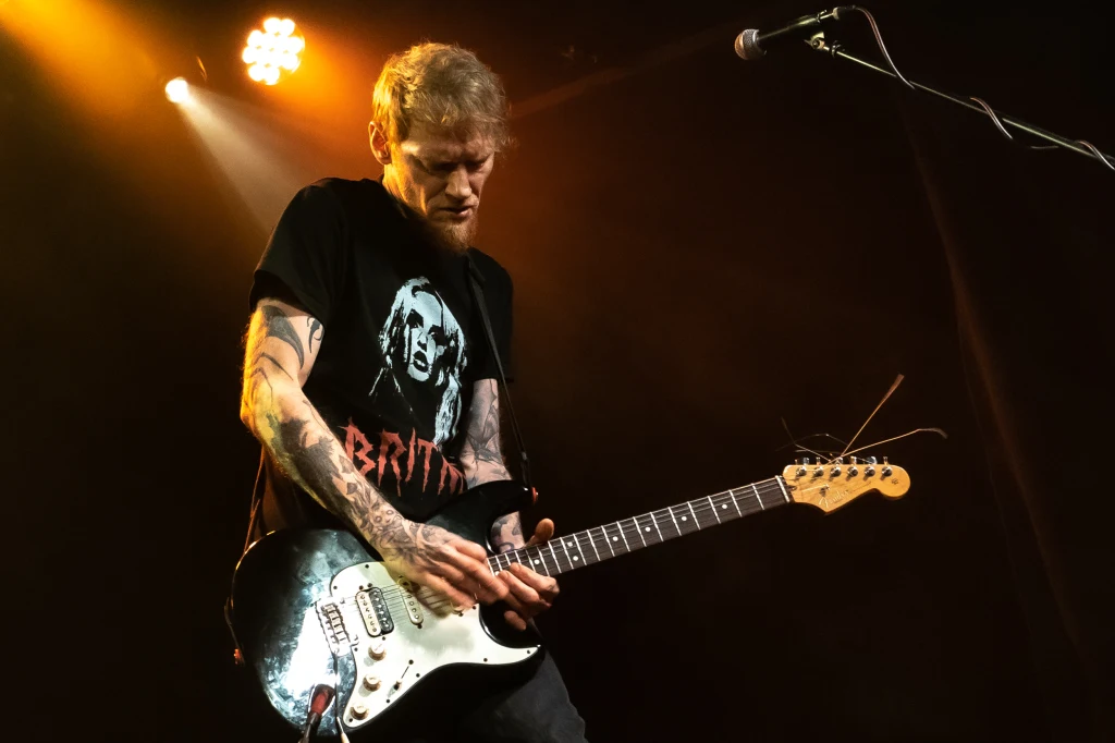 Dawid Karpiuk (Dziwna Wiosna) na koncercie w Krakowie Gitarzysta grający na elektrycznej gitarze podczas koncertu, ubrany w czarną koszulkę z nadrukiem, oświetlony ciepłym światłem scenicznym, na tle mikrofonu.