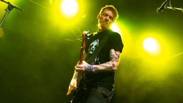 Muzyk grający na gitarze elektrycznej na scenie, oświetlony intensywnym żółtym światłem. Tatuaże na rękach artysty oraz czarna koszulka z nadrukiem.