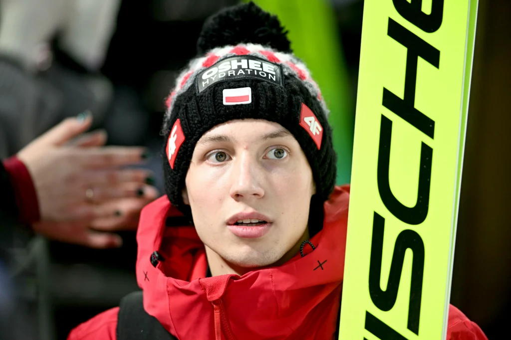 Młody mężczyzna ubrany w zimową czapkę z logo sponsora i polską flagą oraz sportową kurtkę w intensywnym czerwonym kolorze, trzyma w ręku narty do skoków narciarskich. W tle widoczna jest rozmazana sylwetka innej osoby.