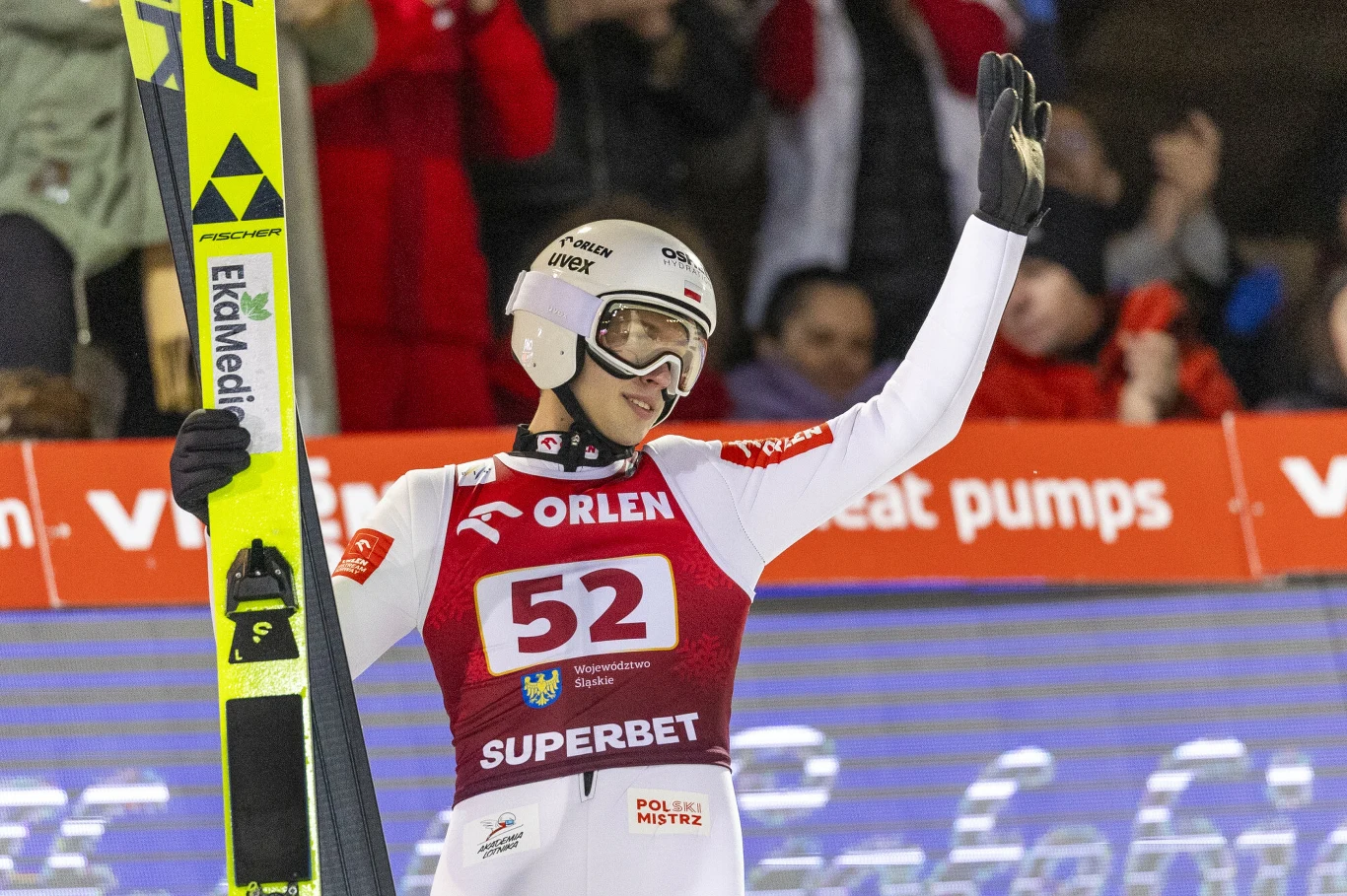 Kacper Tomasiak Skoczek narciarski ubrany w kombinezon startowy z numerem 52, kaskiem i goglami unosi rękę w geście pozdrowienia, trzymając narty, wokół widoczny tłum kibiców oraz reklamy partnerów zawodów.
