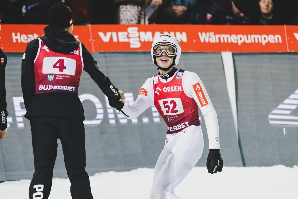 Dwóch skoczków narciarskich ubranych w białe i czarne stroje sportowe ściska sobie dłonie na tle trybun oraz banerów reklamowych. Zawodnik z numerem 52 szeroko uśmiecha się, a atmosfera jest pełna sportowych emocji.