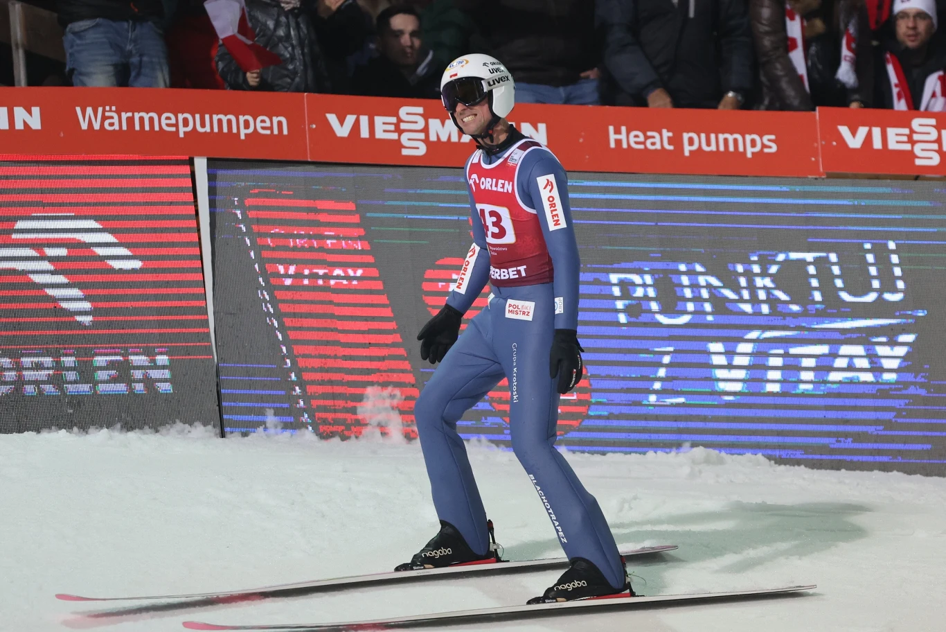 Zawodnik w kombinezonie narciarskim i kasku ląduje na nartach na śnieżnej skoczni, na tle reklam i kibiców, podczas zawodów skoków narciarskich.