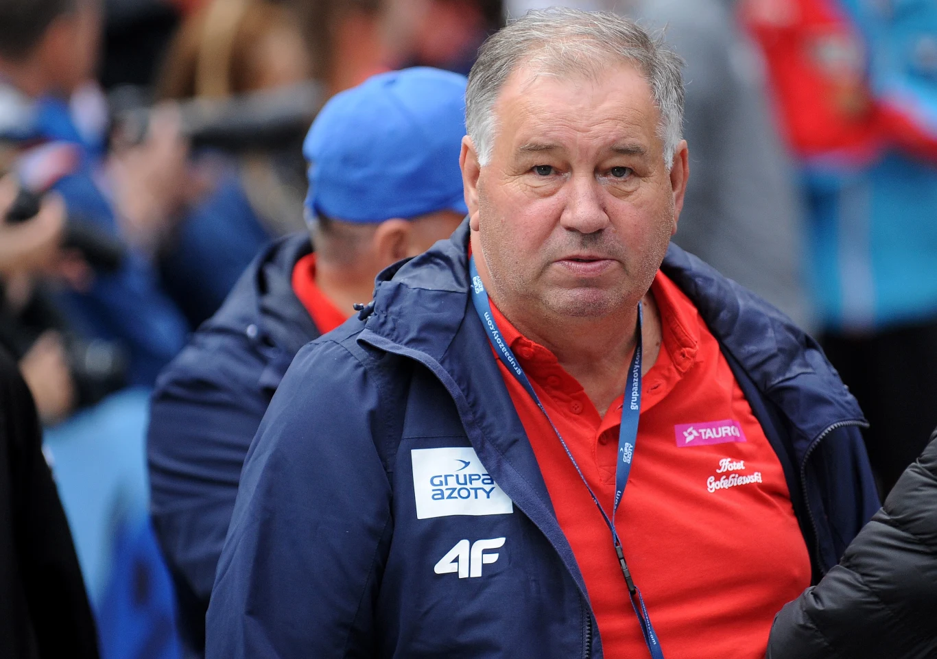 Andrzej Wąsowicz Starszy mężczyzna w sportowej kurtce i czerwonej koszulce z logotypami sponsorów znajduje się na pierwszym planie, patrząc w stronę aparatu. W tle rozmyci ludzie w niebieskich i czerwonych ubraniach, co sugeruje atmosferę wydarzenia sportowego.