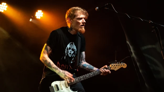 Mężczyzna z tatuażami gra na gitarze elektrycznej podczas koncertu na scenie, ubrany w czarną koszulkę z nadrukiem, oświetlony światłami estradowymi.