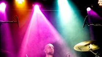 Perkusista grający energicznie na zestawie perkusyjnym w barwnym świetle reflektorów scenicznych, wokół unosi się delikatna mgła tworząca koncertową atmosferę.