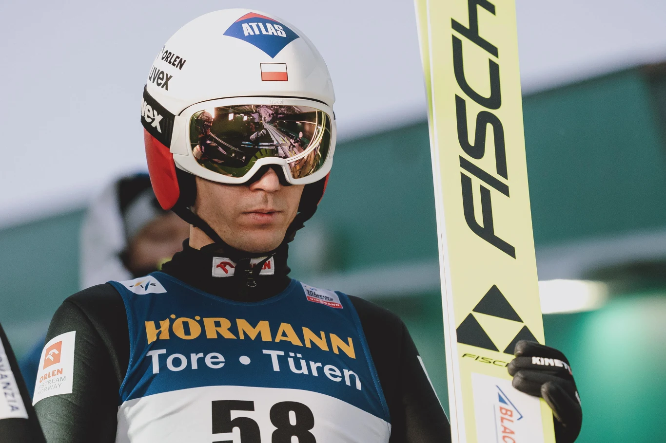 Kamil Stoch Skoczek narciarski w stroju startowym, kasku i goglach, trzymający narty Fischer, przygotowuje się do skoku podczas zawodów. Na piersi numer startowy oraz logo sponsorów.
