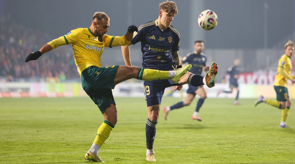 Alan Czerwiński (z lewej) kontra Patryk Makuch Dwóch piłkarzy walczy o piłkę podczas intensywnej akcji na murawie stadionu, w tle widoczni inni zawodnicy oraz rozmyty stadion z publicznością.