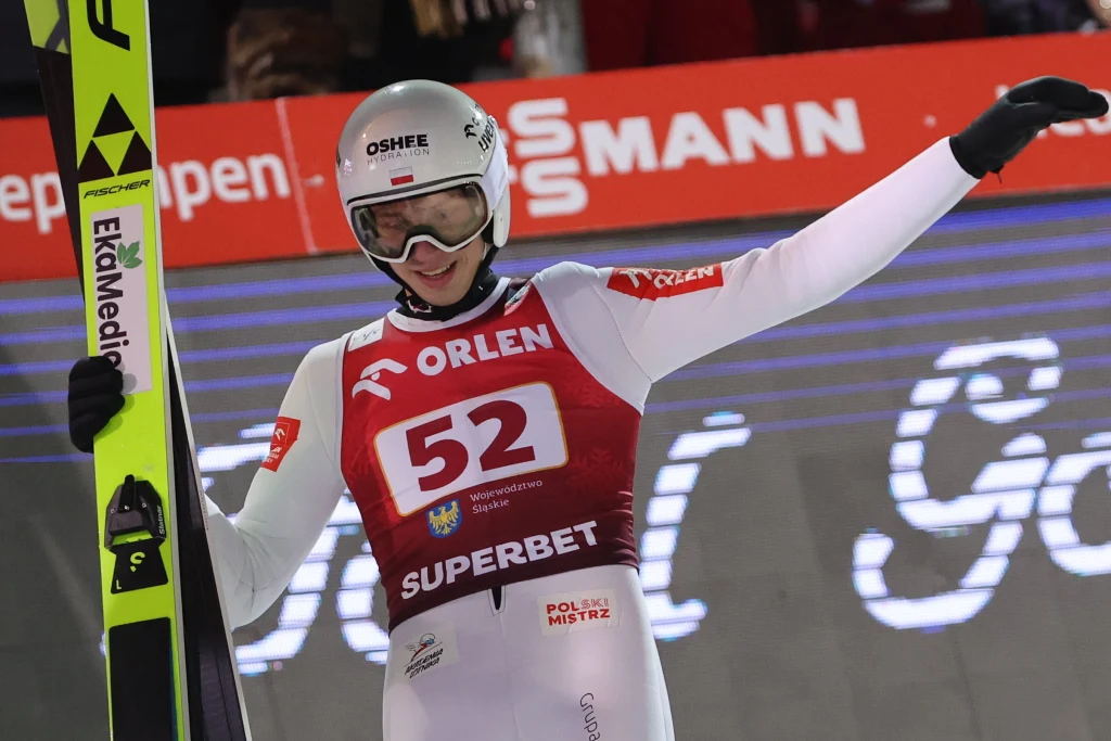 Kacper Tomasiak Sportowiec ubrany w kombinezon do skoków narciarskich z numerem startowym 52 unosi rękę w geście radości trzymając narty, na tle reklamy sponsorów i tłumu kibiców.