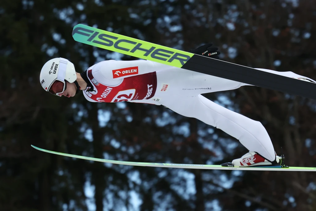 Kacper Tomasiak Skoczek narciarski ubrany w biały kombinezon oraz kask podczas lotu na nartach, w tle rozmyte drzewa, widoczne logo sponsora na nartach oraz numer startowy na piersi.