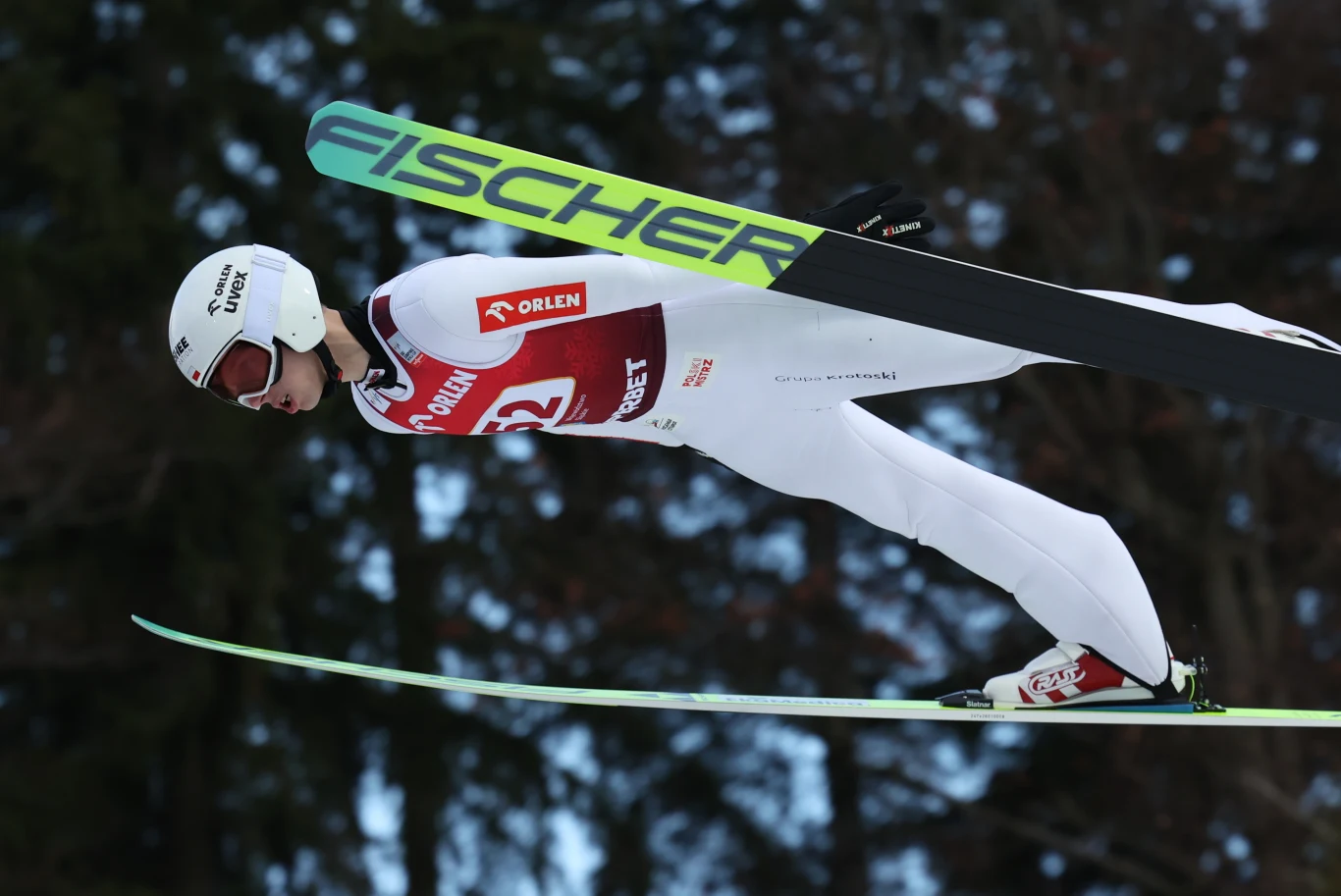 Skoczek narciarski ubrany w biały kombinezon oraz kask podczas lotu na nartach, w tle rozmyte drzewa, widoczne logo sponsora na nartach oraz numer startowy na piersi.