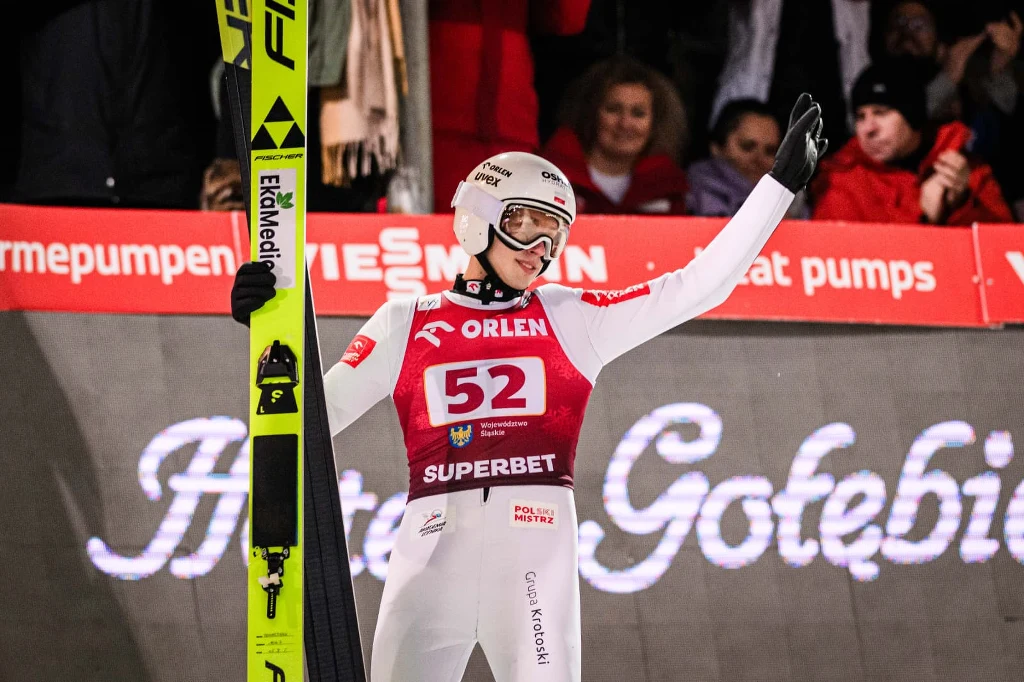 Kacper Tomasiak Zawodnik skoków narciarskich w pełnym stroju sportowym stoi z uniesioną ręką, trzymając narty. W tle widoczna publiczność oraz bannery reklamowe.