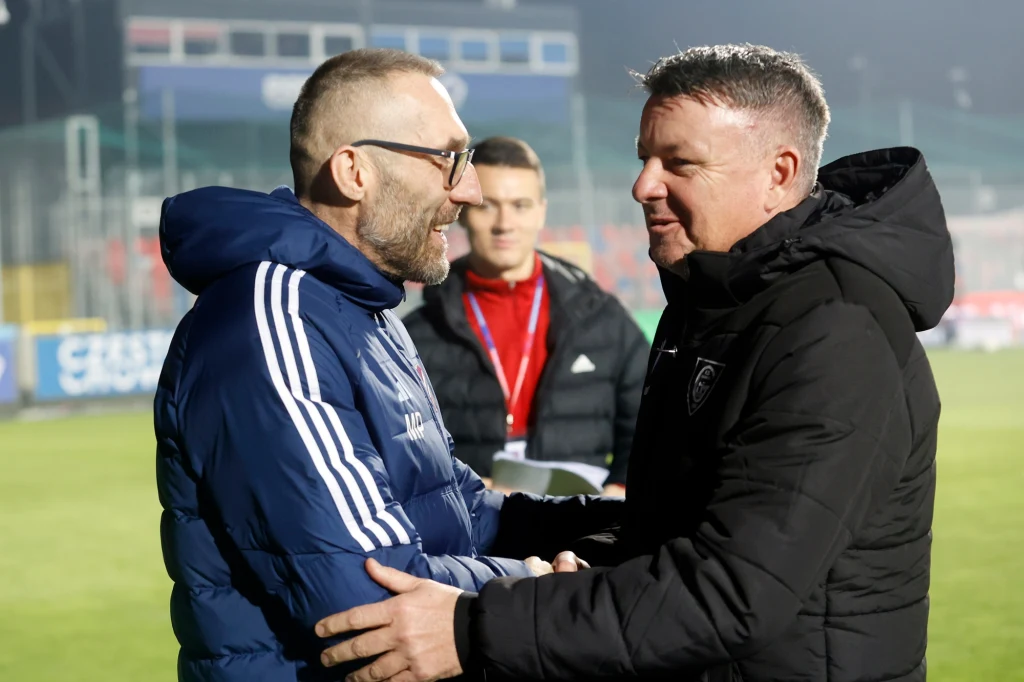 Marek Papszun (z lewej) wita się z Rafałem Górakiem, opiekunem "Gieksy" Dwóch mężczyzn w sportowych kurtkach uśmiecha się i ściska dłonie na tle stadionu piłkarskiego, w tle trzeci mężczyzna obserwuje sytuację.