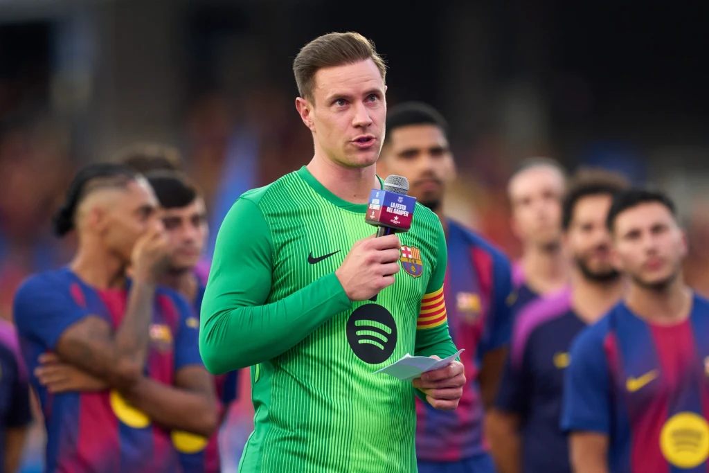 Marc-Andre ter Stegen Bramkarz w zielonym stroju z opaską kapitańską trzyma mikrofon i kartkę podczas przemówienia, w tle stoją piłkarze drużyny w niebiesko-bordowych koszulkach z logo sponsora, skupieni i słuchający.