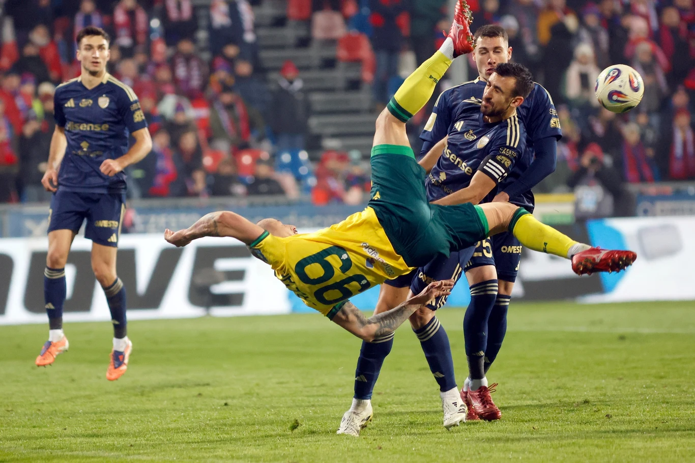 Zawodnik w żółtej koszulce wykonuje przewrotkę podczas meczu piłki nożnej, otoczony przez innych piłkarzy w granatowych strojach, tło stanowi trybuna z kibicami.