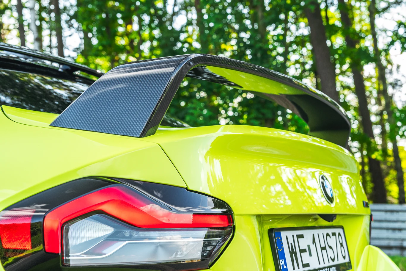 Gdy się dobrze przyjrzeć, okaże się, że testowane M2 ma aż trzy tylne spojlery. Jeden zintegrowany jest z klapą bagażnika, drugi (karbonowy) góruje nad nią, a nad tylną szybą umieszczono trzeci Jaskrawożółty samochód sportowy z widocznym tylnym skrzydłem z włókna węglowego, charakterystycznym agresywnym tylnym światłem oraz polską tablicą rejestracyjną, na tle gęstego, zielonego lasu.