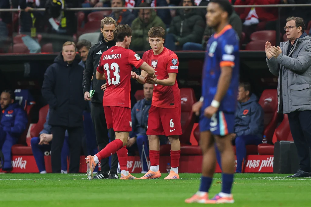 Dwóch piłkarzy w czerwonych strojach reprezentacji Polski wymienia się na murawie podczas meczu. W tle ławka trenerska oraz widoczni członkowie sztabu szkoleniowego. Na pierwszym planie zawodnik drużyny przeciwnej w niebiesko-pomarańczowym stroju.