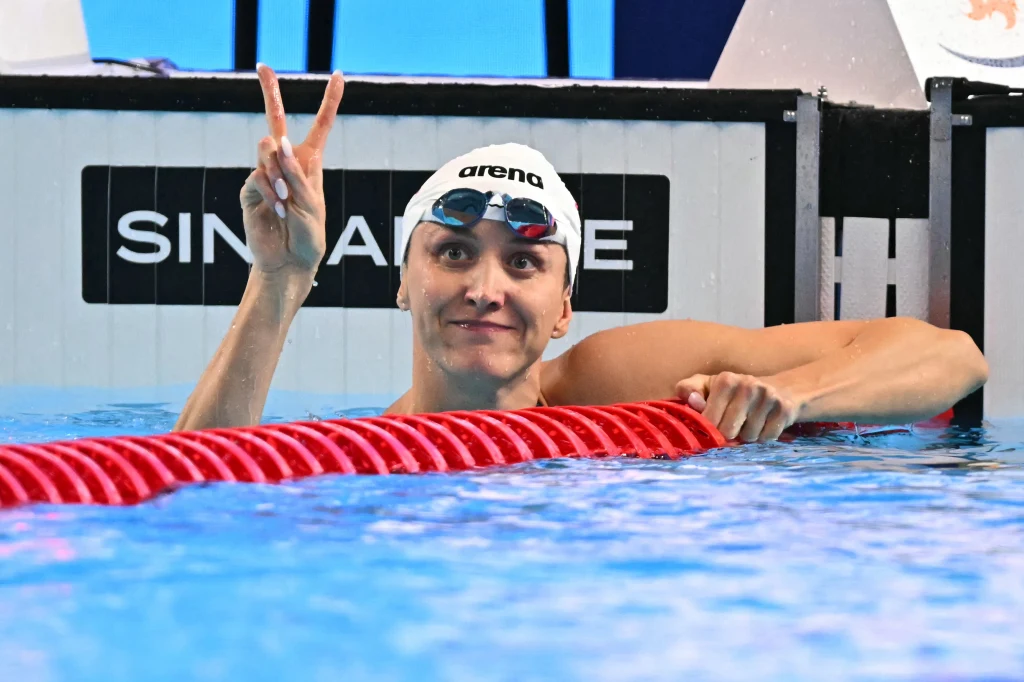 Katarzyna Wasick ze złotym medalem na mistrzostwach Europy w Lublinie Pływak unosi się przy krawędzi basenu, opierając rękę o czerwony tor, pokazując znak pokoju dwoma palcami. Ma na sobie czepek, okulary pływackie i uśmiecha się do aparatu.